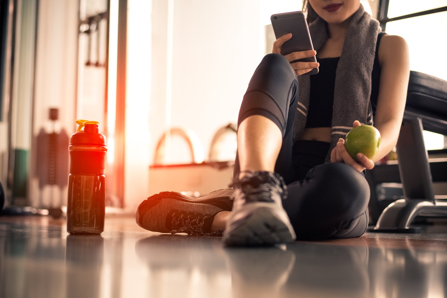 A woman checking her phone while resting on the gym floor with a water bottle nearby