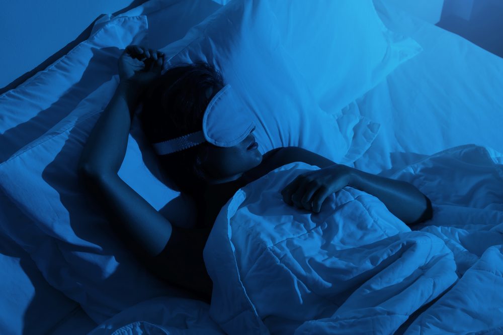 A woman asleep under blue-toned bedding while wearing a sleep mask