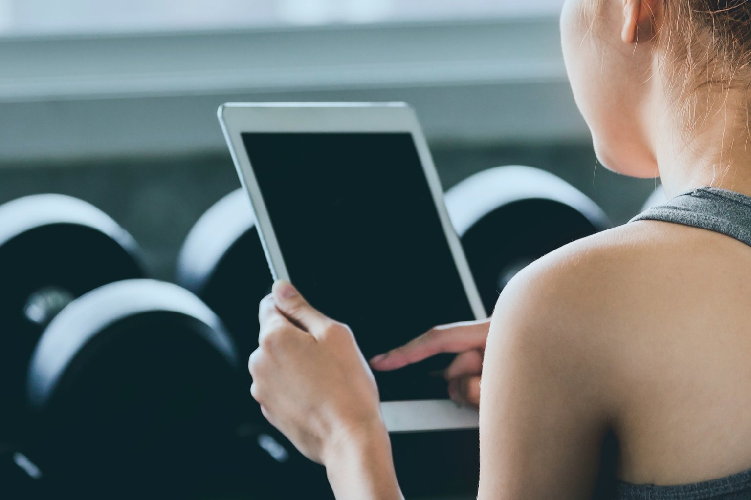 A woman in the gym looking up an exercise on a tablet near a dumbbell rack