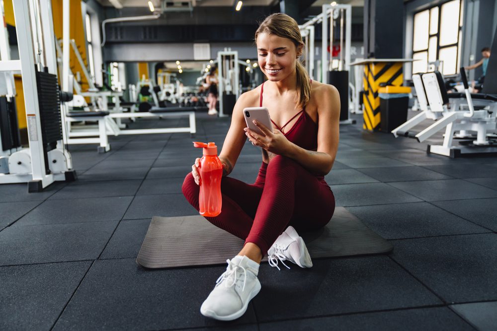 A woman sitting on a gym mat checking her phone after finding an exercise