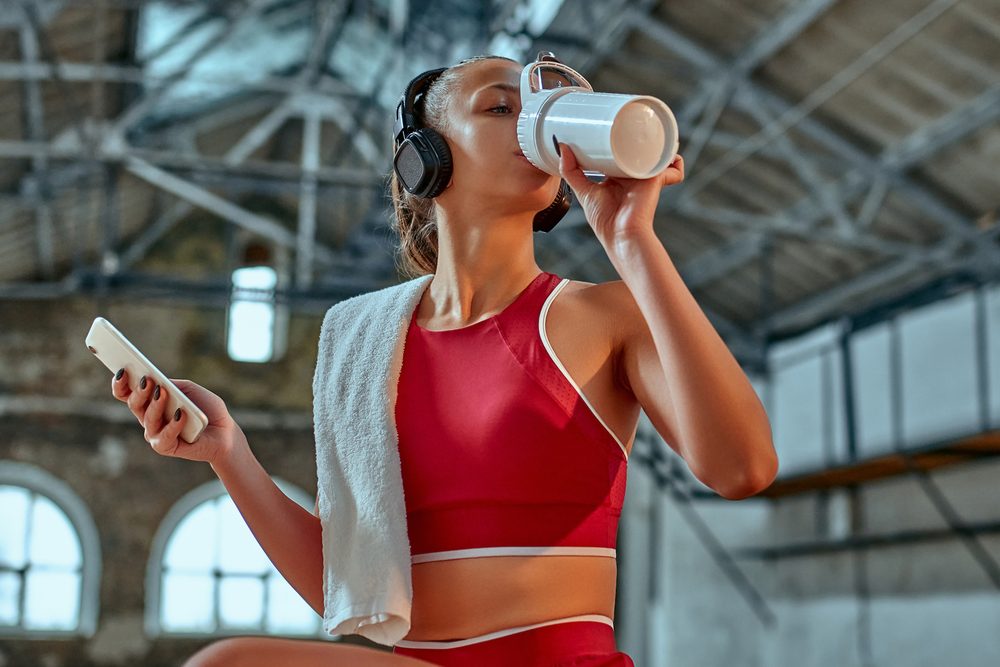 A woman drinking from a water bottle after completing a workout, headphones on