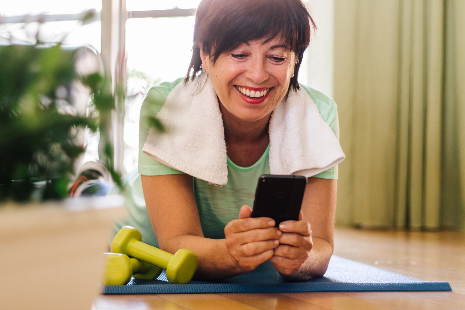 A woman smiling while checking her phone on a yoga mat at home, surrounded by light dumbbells