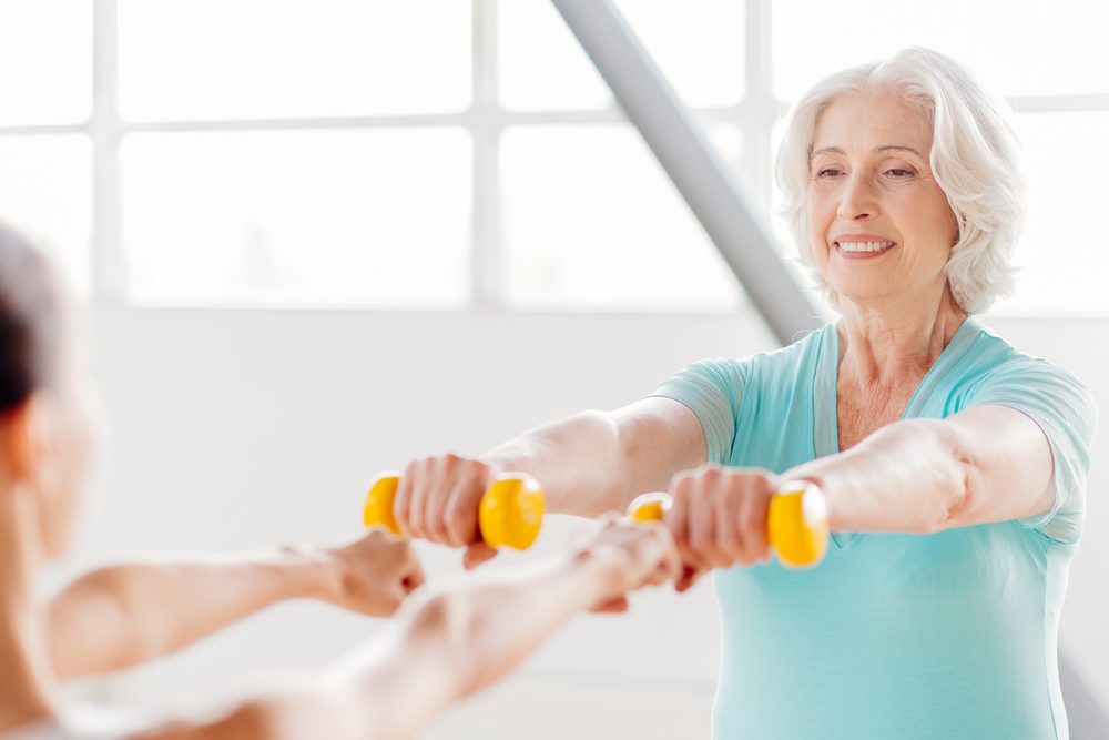 An older woman training with dumbbells alongside another person in a bright gym